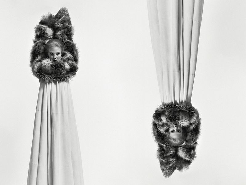 Monochrome fashion editorial image showing a model standing upright and an inverted model hanging above, wearing fur-trimmed garments.