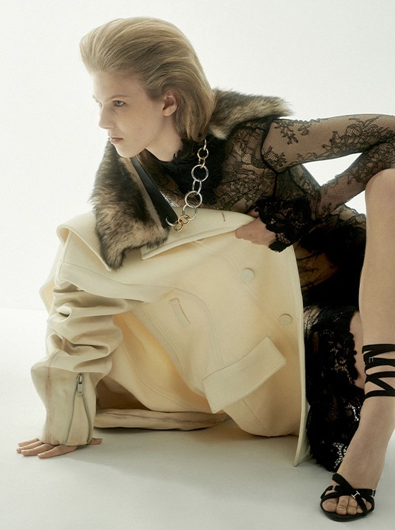 Fashion model poses kneeling in a lace top, fur-collared coat, and sandals on a white background in a studio fashion shot.
