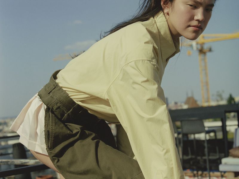 Outdoor fashion shot of a young adult Asian woman wearing a light-yellow collared shirt and green shorts, posing on a rooftop on a sunny day.