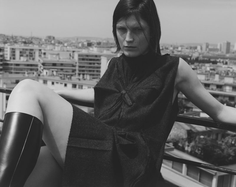 Woman in a dark dress and black boots sitting on a railing with a cityscape in the background, creating a monochrome fashion portrait.