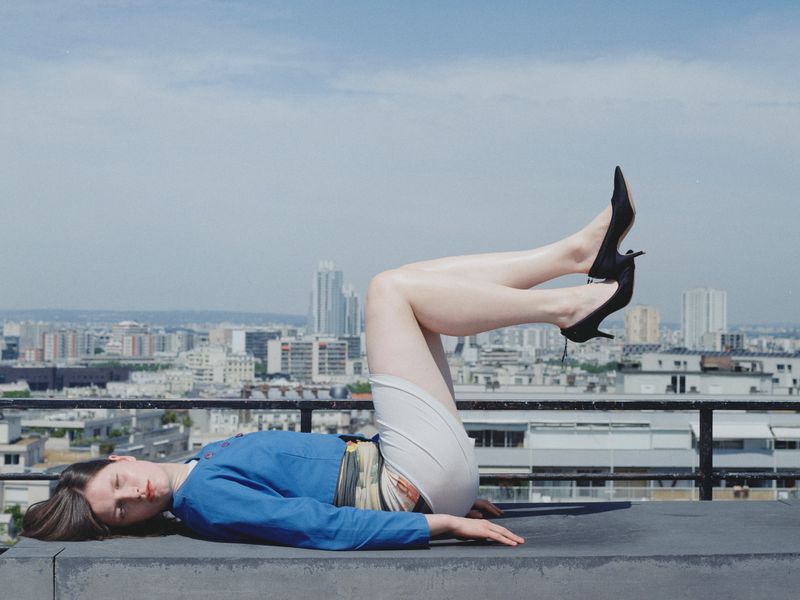 Adult woman lounges on a rooftop with her legs up in black heels, with an urban city skyline and blue sky in the background.