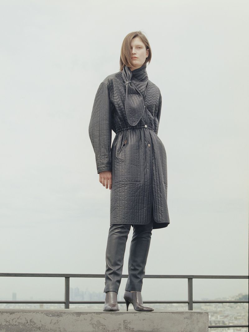 Model in a quilted dark midi coat and heeled ankle boots standing on a concrete rooftop ledge against an overcast sky.