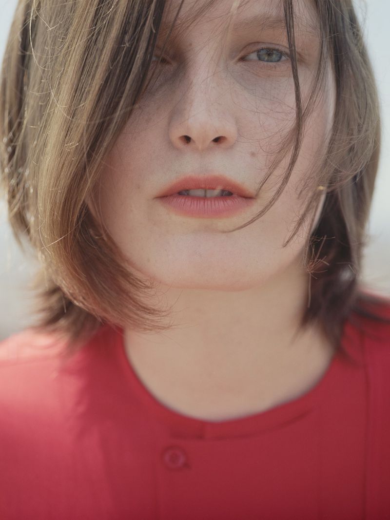 Close-up portrait of a young adult female with fair skin and shoulder-length brown hair wearing a red blouse, shot outdoors.