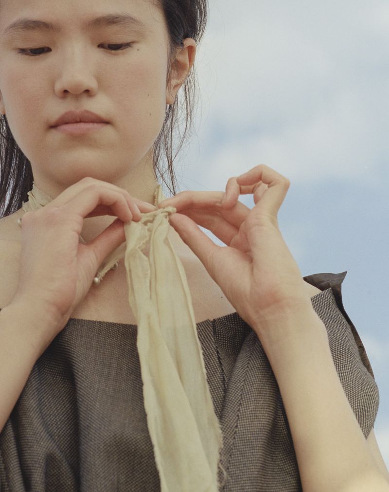 A young adult woman ties a silky off-white scarf against a backdrop of a bright blue sky with light clouds, creating a serene, fashion-focused image.