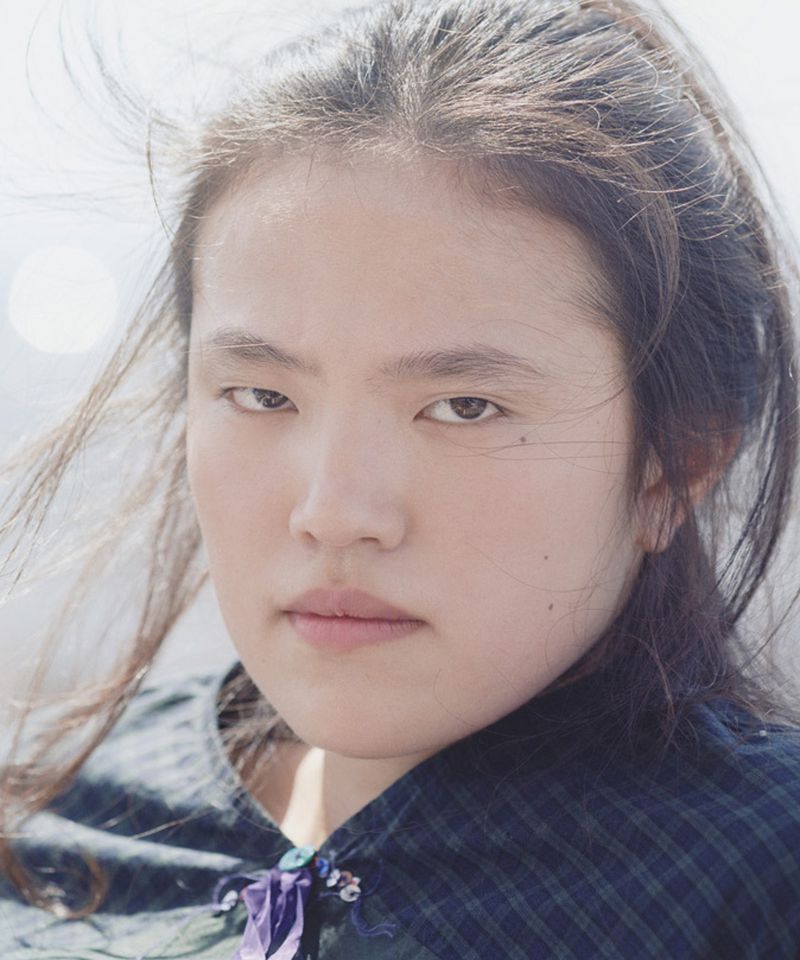 Portrait of a woman with windblown hair, wearing a blue plaid shirt with ribbon detail, against a bright, blurred outdoor background.