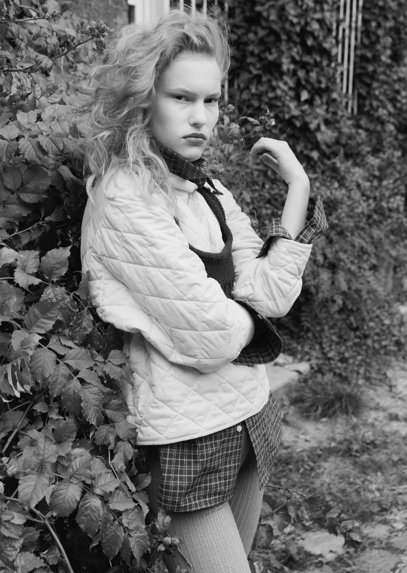 Monochrome portrait of a young woman with curly hair wearing a quilted jacket and plaid shirt, leaning against a leafy wall.