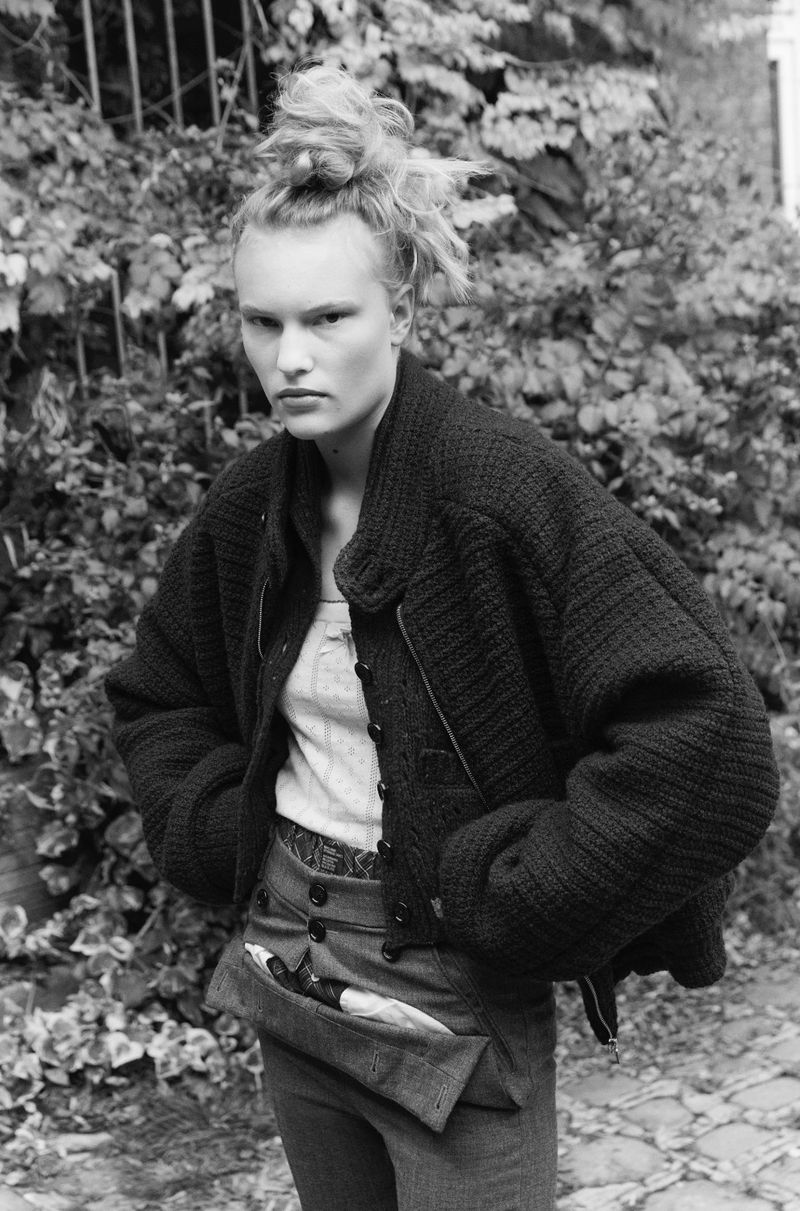 A woman poses in a jacket and unique pants in a leafy outdoor setting, captured in a black and white photograph with a somber mood.
