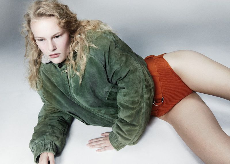 Woman lies on a white surface wearing a green jacket and orange shorts, showcasing fashion style in a studio environment with soft lighting.