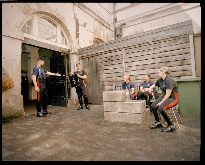 Group of people in uniform sit outdoors near stone benches with buildings in the background in a medium eye-level shot.