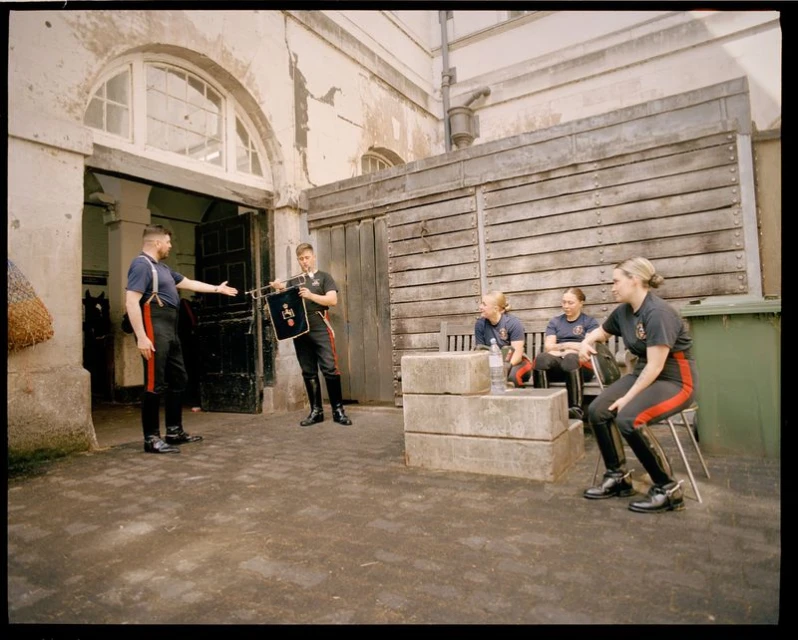 Group of people in uniform sit outdoors near stone benches with buildings in the background in a medium eye-level shot.