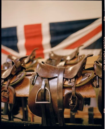 Several brown leather horse saddles are displayed on a wooden rack, with a British Union Jack flag hanging in the background.