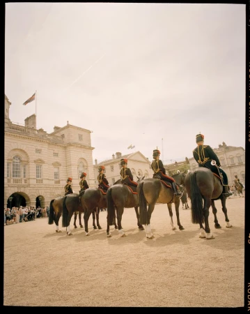 Mounted members of the Household Cavalry in ceremonial uniform on horseback in front of a light-colored building with a flag on the roof.