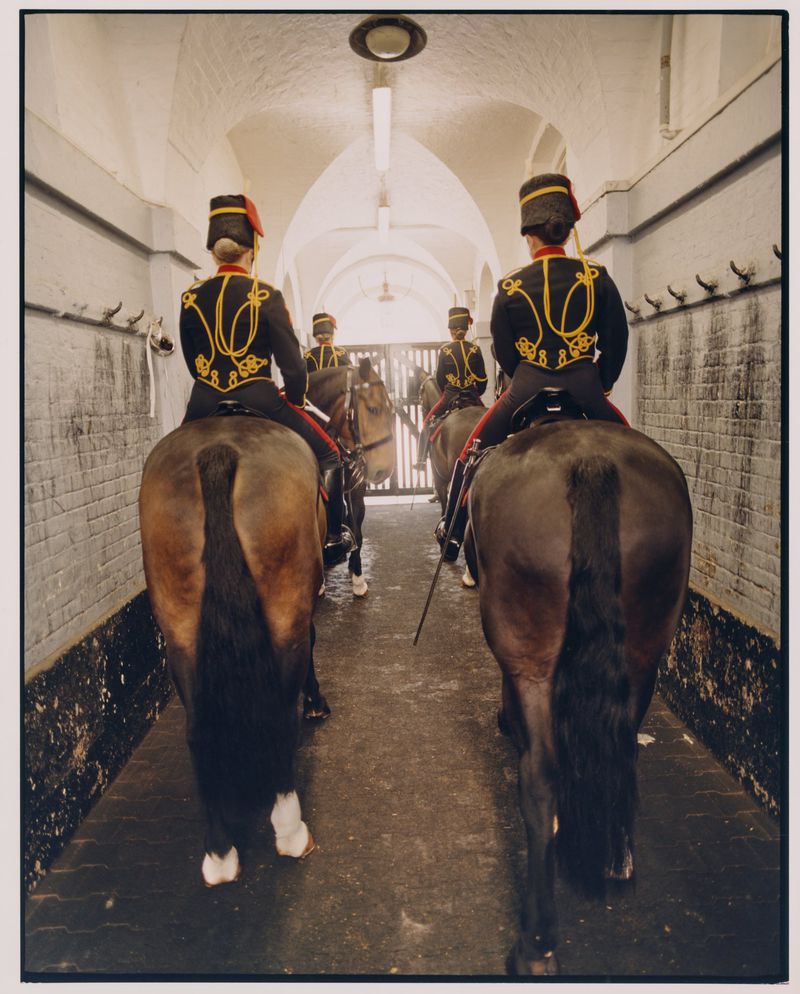 Mounted royal guards in formal attire ride horses in a tunnel with a vaulted ceiling, creating a symmetrical composition.