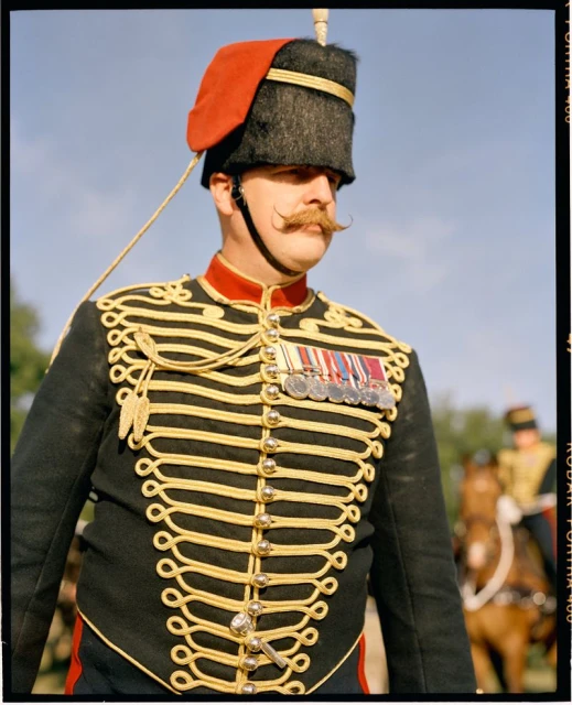 Man in ornate military uniform and red-topped hat with a handlebar mustache. Medals displayed on coat. Outdoor setting.