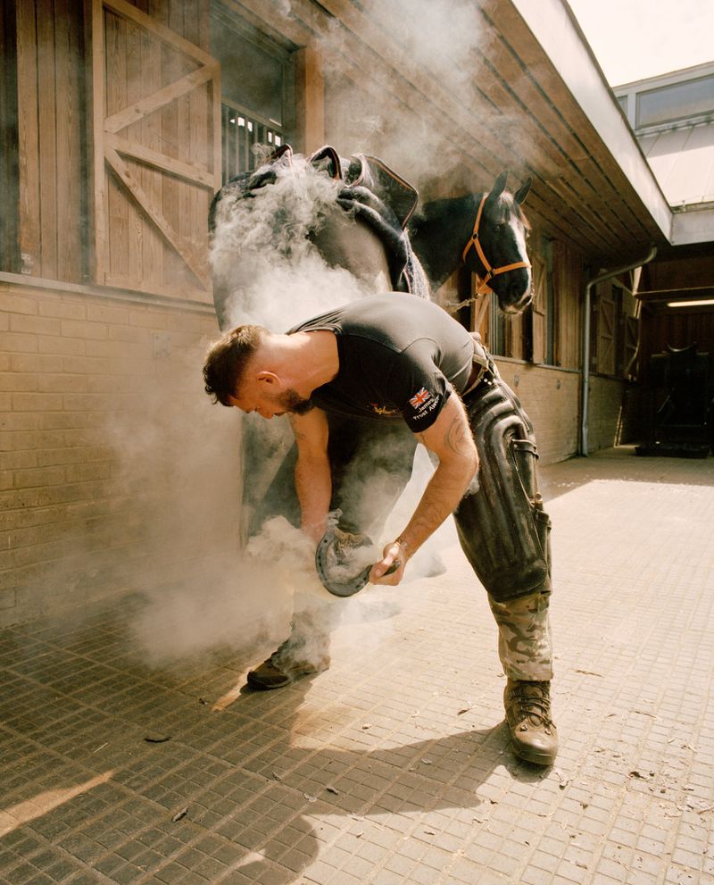 Adult male farrier fitting horseshoe on a horse with smoke. Horse in the background, standing outdoors in front of a barn.