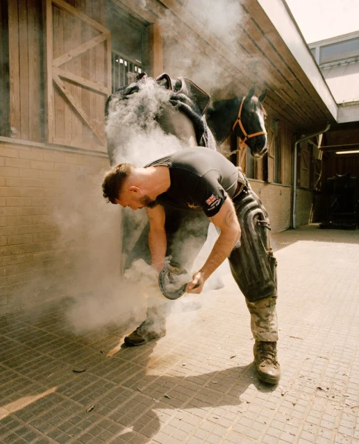 Adult male farrier fitting horseshoe on a horse with smoke. Horse in the background, standing outdoors in front of a barn.