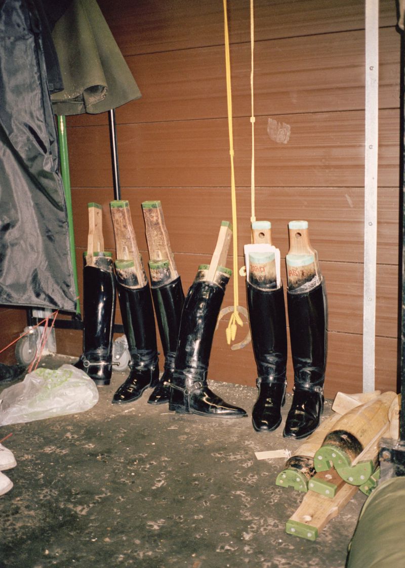 Several pairs of shiny black riding boots are lined up indoors, some with wooden boot shapers inside. One has a yellow pull strap.