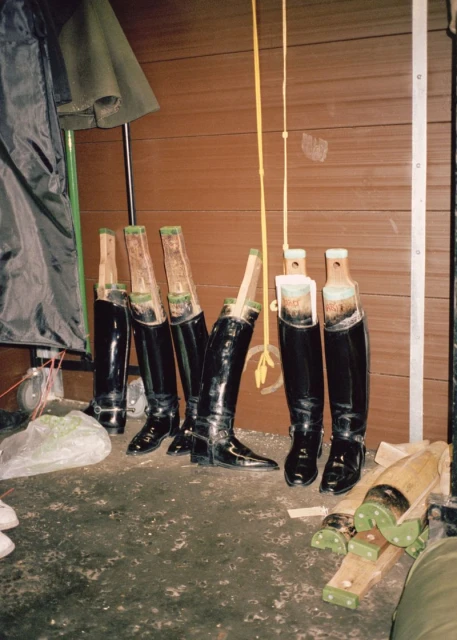 Several pairs of shiny black riding boots are lined up indoors, some with wooden boot shapers inside. One has a yellow pull strap.