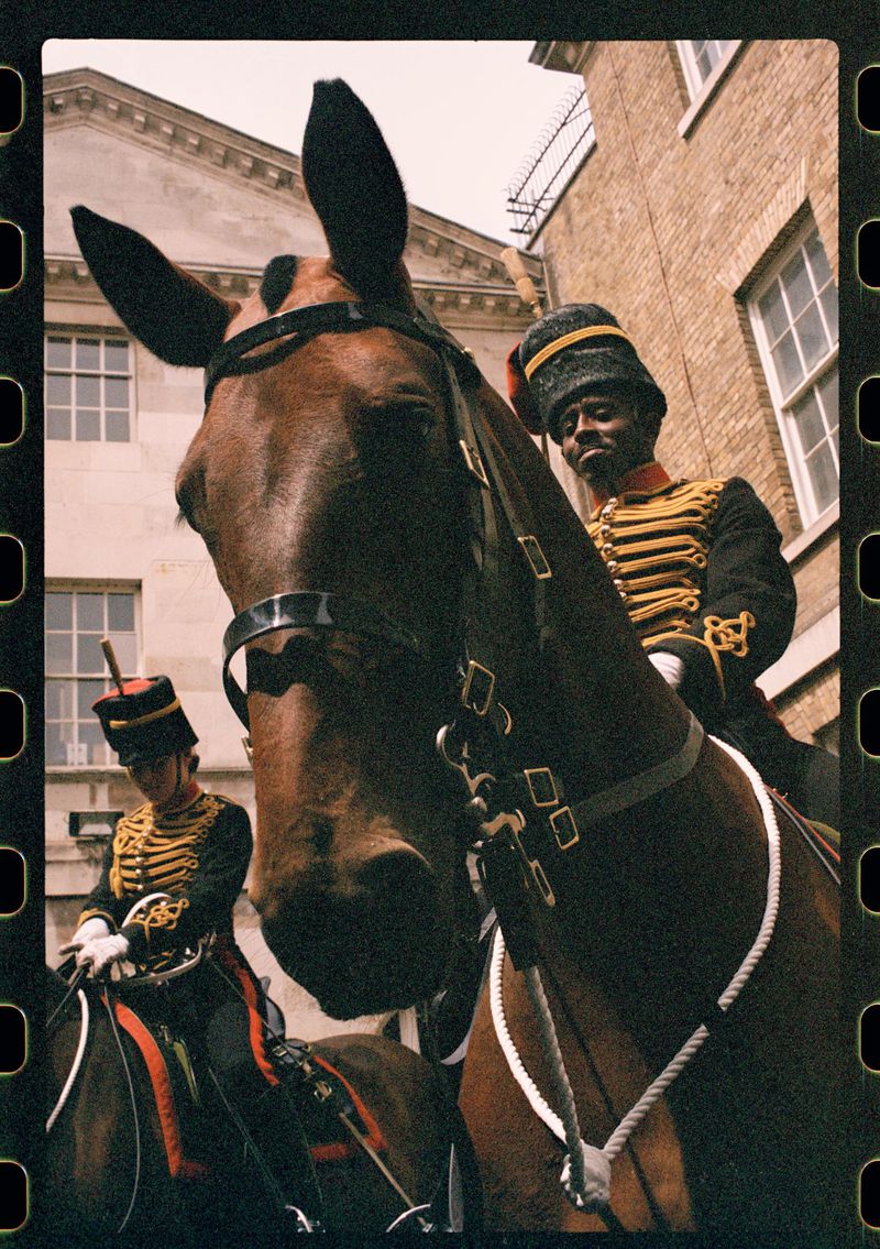 Two mounted Household Cavalry soldiers in ceremonial uniform, one in the foreground, with a government building in the background.