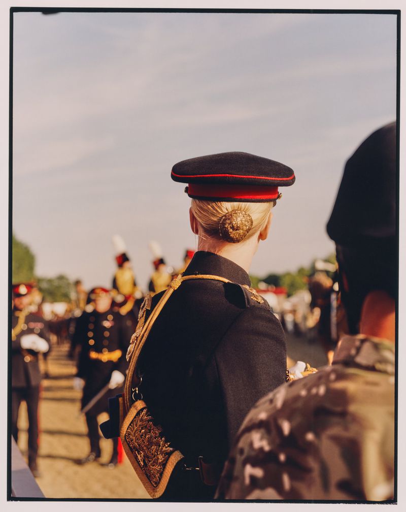 Rear view of a person in military dress uniform with a hat in a crowd, outdoors on a sunny day, at eye-level angle, wearing an intricate outfit.