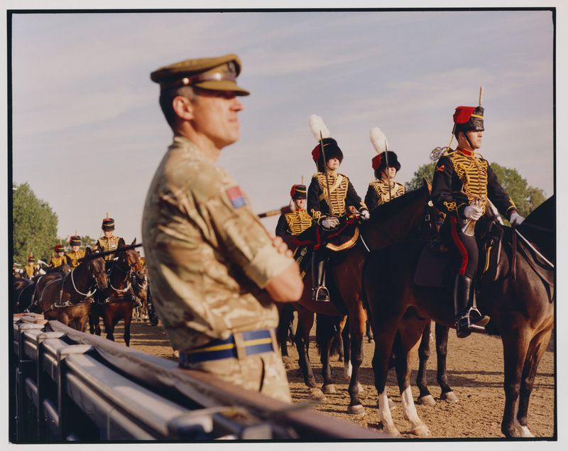 British Army ceremonial unit on horseback. Soldiers wearing traditional uniforms with red jackets and plumed helmets. Man in camouflage uniform.