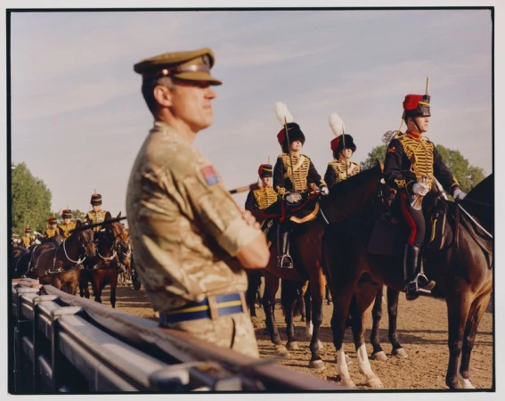 British Army ceremonial unit on horseback. Soldiers wearing traditional uniforms with red jackets and plumed helmets. Man in camouflage uniform.