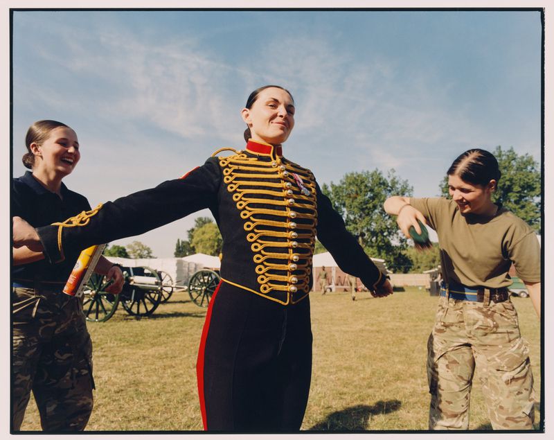 Three women in a grassy field, one in a military uniform with gold trim, the others in camouflage, with trees and tents in the background.