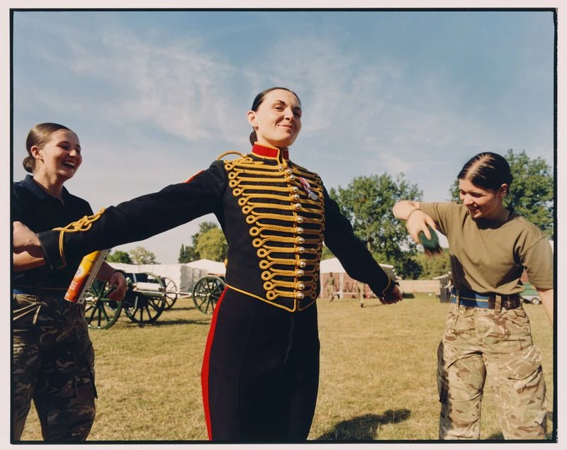 Three women in a grassy field, one in a military uniform with gold trim, the others in camouflage, with trees and tents in the background.