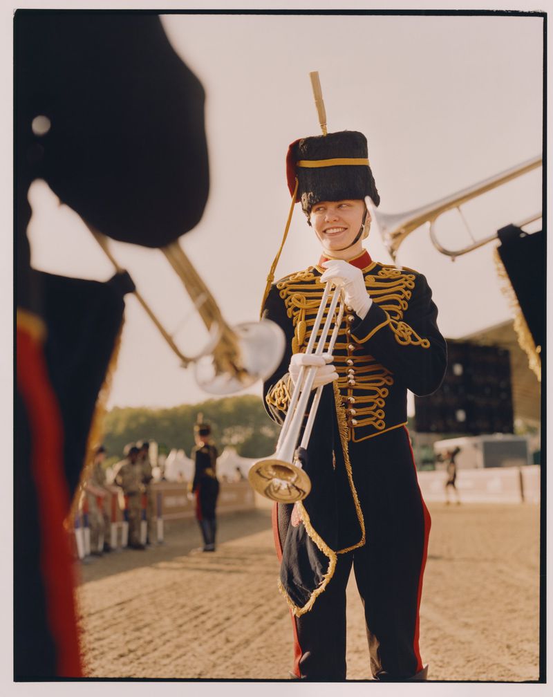 Smiling musician in uniform holds trumpet, captured in medium shot, outdoors with other musicians in the background.