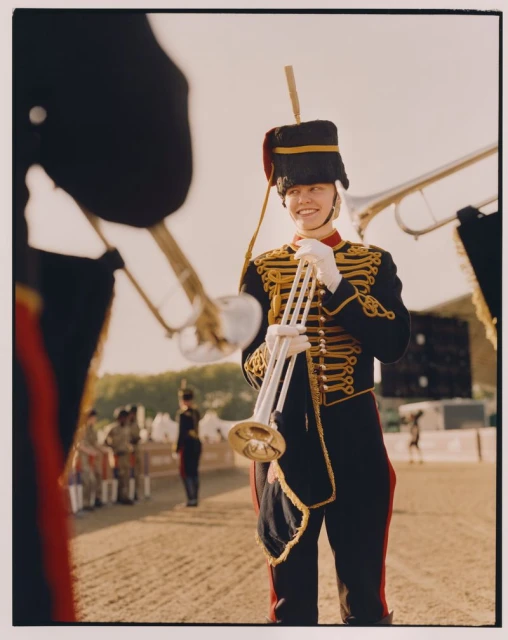 Smiling musician in uniform holds trumpet, captured in medium shot, outdoors with other musicians in the background. 
