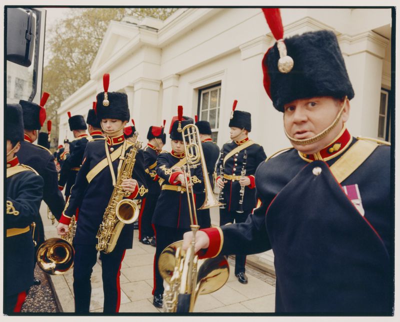 Marching band wearing military uniforms playing instruments outdoors in front of a white building, medium shot.
