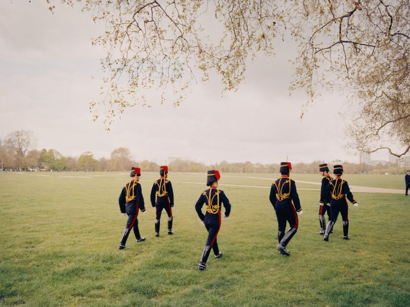 Group of guards in black and red uniforms marching on a green lawn under trees against a light sky, creating a formal scene.