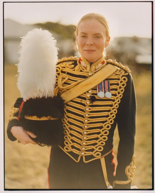 Portrait of a woman in a military uniform holding a white plume hat, standing outdoors with a blurred landscape background during daytime.