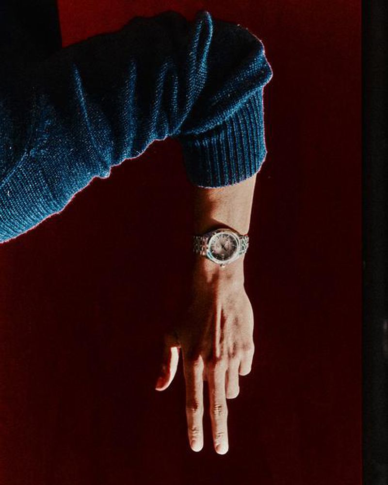 Close-up of an arm with a silver watch, fingers pointing down, set against a solid red background and a person wearing a blue sweater.