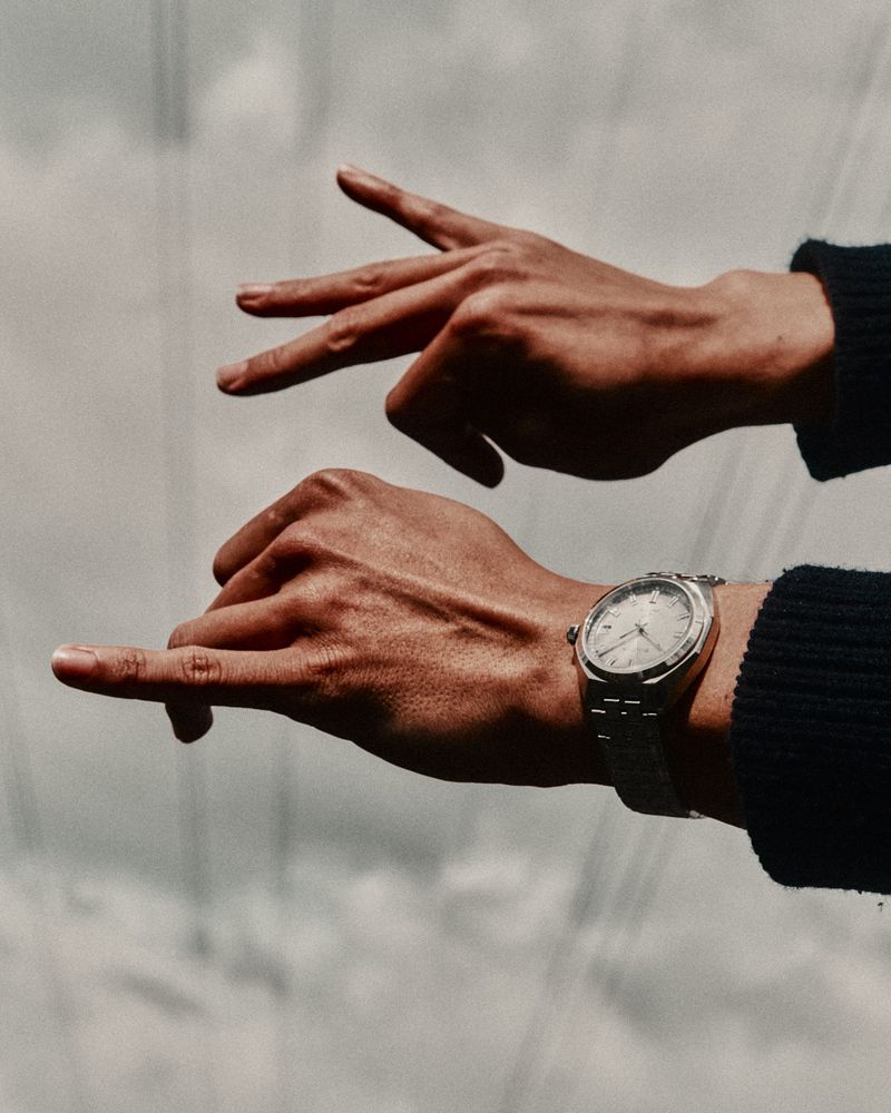 Close-up shot of two hands with a wristwatch against a cloudy sky backdrop. One hand is open, the other is making a gesture.