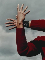 Close-up of hands raised in an expressive gesture wearing a black wristwatch and a crimson knit sweater against an overcast sky.