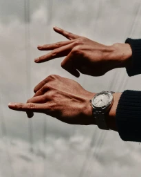 Close-up of hands gesturing against a pale cloudy sky, with one wrist wearing a silver steel watch and dark knit cuff.