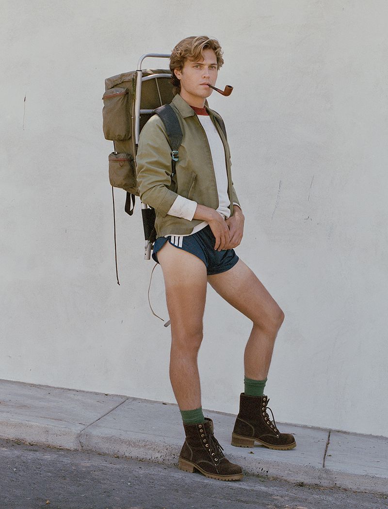 An adult male stands with a backpacking frame and brown boots, smoking a pipe against a textured white wall in an outdoor environment.