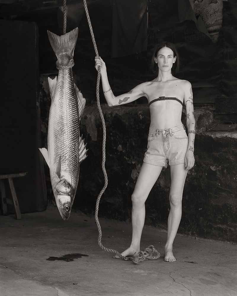Monochrome portrait of a barefoot woman in denim shorts holding a rope tied to a large hanging fish in a rustic interior.