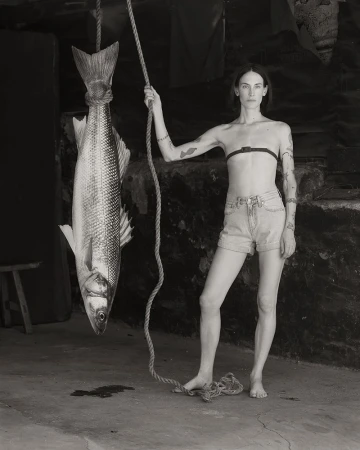 Monochrome portrait of a barefoot woman in denim shorts holding a rope tied to a large hanging fish in a rustic interior.