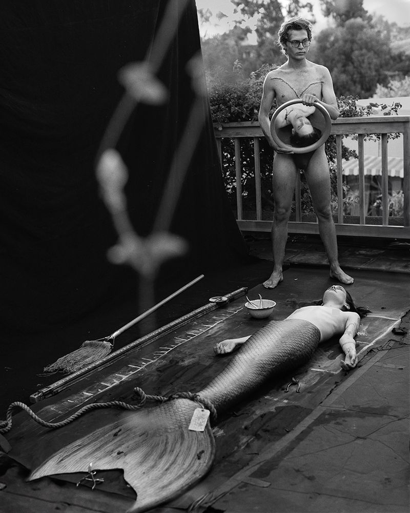 Black and white image featuring a human 'mermaid' lying on a black surface, with a man holding a mirror reflecting a face, outdoors.