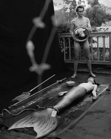 Black and white image featuring a human 'mermaid' lying on a black surface, with a man holding a mirror reflecting a face, outdoors.