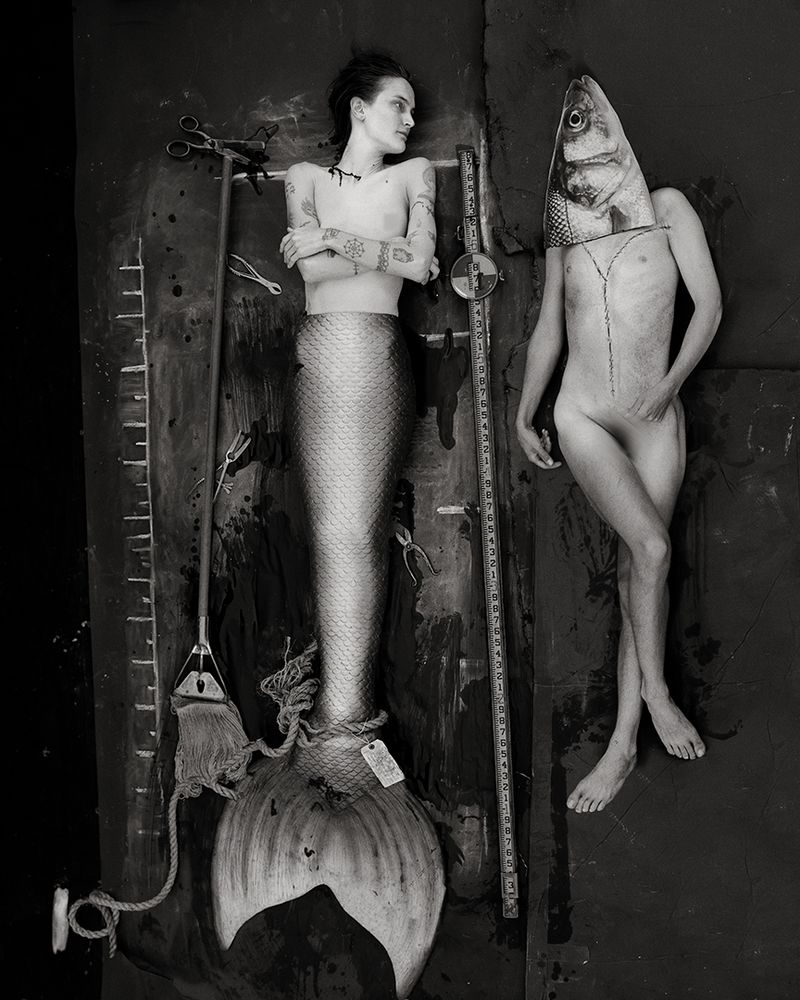 Monochrome image of a tattooed woman as a mermaid lying on a dark surface with surgical tools and a person with a fish head.
