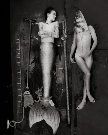Monochrome image of a tattooed woman as a mermaid lying on a dark surface with surgical tools and a person with a fish head.