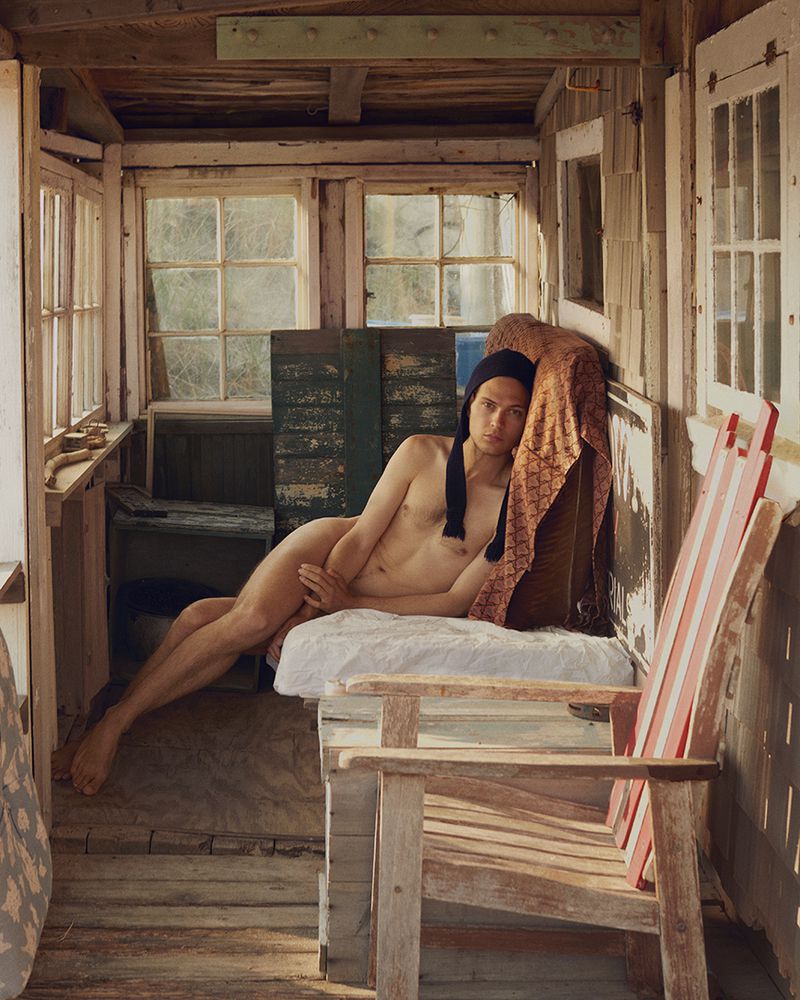 Nude male reclines on daybed in rustic porch setting, wearing a cap, natural light casting shadows through the windows, weathered textures.