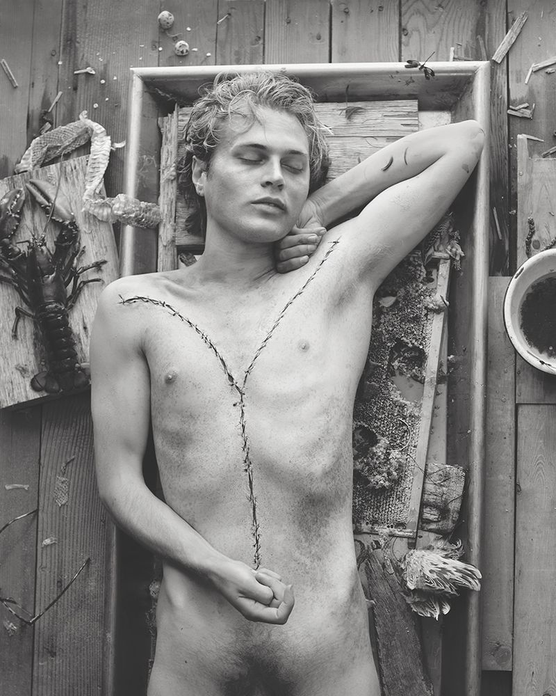 Monochrome portrait of a shirtless male model with closed eyes lying in a box frame adorned with barbed wire and rustic elements.