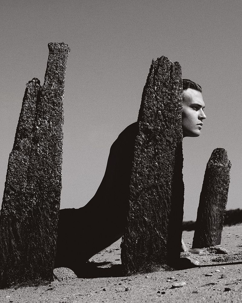 Monochrome portrait of adult male partially obscured by stone pillars in a sandy landscape with a clear sky background.