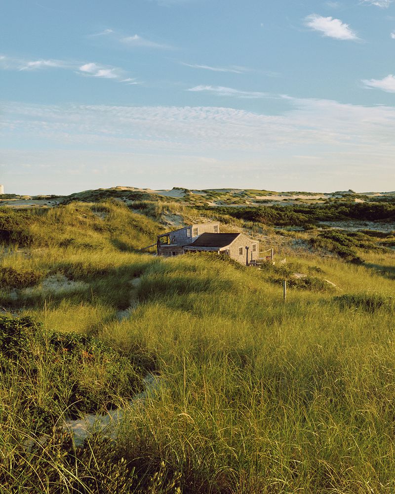 Exterior of a gray house nestled among grassy dunes under a clear blue sky with scattered clouds, bathed in soft, natural light.
