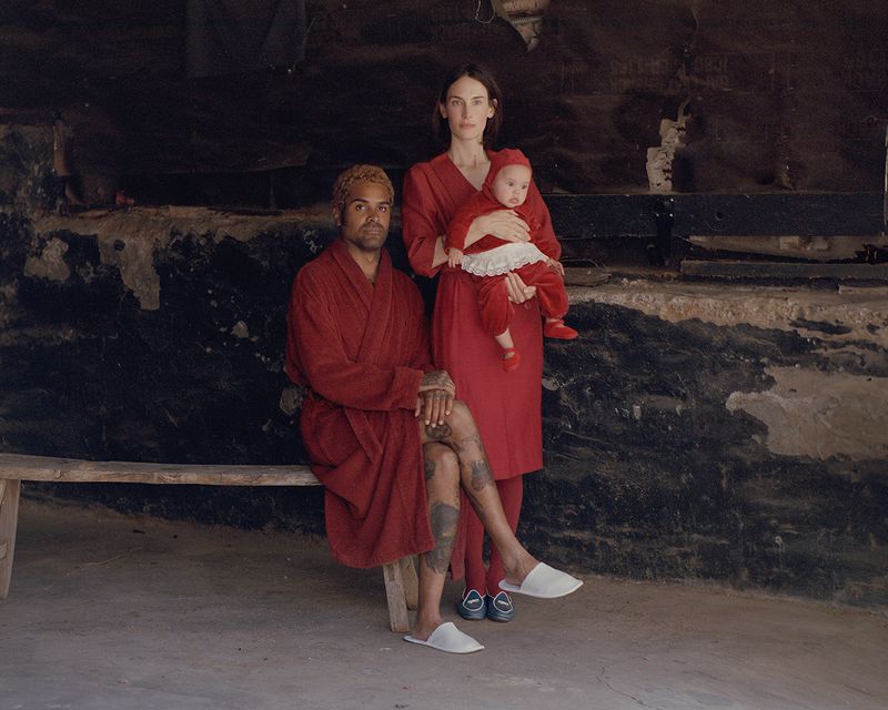 Portrait of a biracial family of three in red outfits, seated on a wooden bench against a textured, dark background inside.