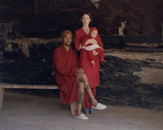 Portrait of a biracial family of three in red outfits, seated on a wooden bench against a textured, dark background inside.